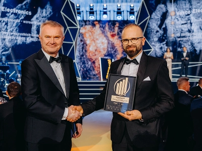 A medium-long shot of two men in formal black-tie attire shaking hands. The man on the right Maciej Góralski holds a framed award with a gold emblem, identifying it as the 