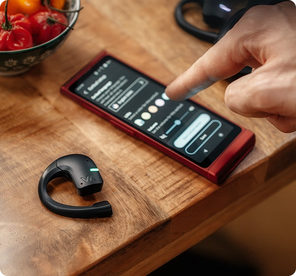A close-up of a person's finger adjusting preferences like color and volume on the screen of a red Vasco Translator V4 device. Next to it on the wooden table rests a black Vasco Tranlator E1 earbud with a glowing green LED indicator, with a bowl of red peppers visible in the background. 