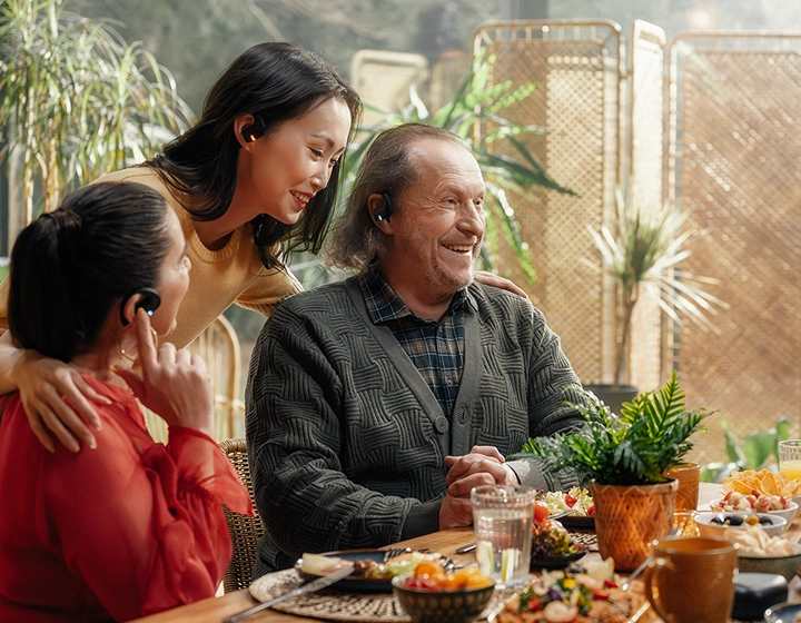 A heartwarming multi-generational family gathering around a dining table filled with food. An older smiling man in a grey cardigan and two women are interacting happily in a sunlit room full of plants. All three individuals are wearing black Vasco Translation E1 earbuds. 