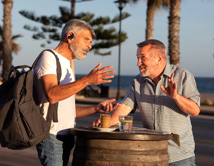 Two men are standing by an outdoor table and having a conversation. One of them is wearing a backpack, and both are using Vasco Translator E1 earbuds. 