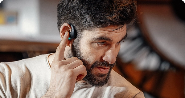 A close-up of a bearded man tapping a black Vasco translation earbud on his ear with his index finger, demonstrating its intuitive touch controls. A small, bright blue LED light is illuminated on the device. 