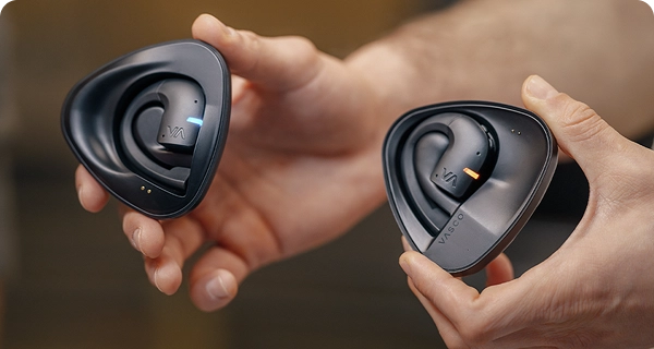 A close-up of hands holding two separate black charging cases with Vasco translation earbuds. The image highlights how to differentiate between the pair, showing one earbud with a blue LED light and the other with an orange LED light for different speakers. 