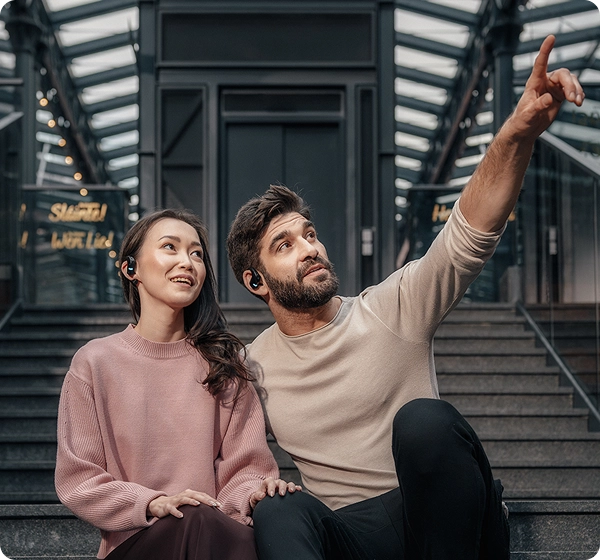 A man and a woman sitting on concrete steps inside a modern architectural structure with a glass canopy. The man points upward with his hand while both wear black Vasco Translation E1 earbuds and look in the direction of his gesture. 