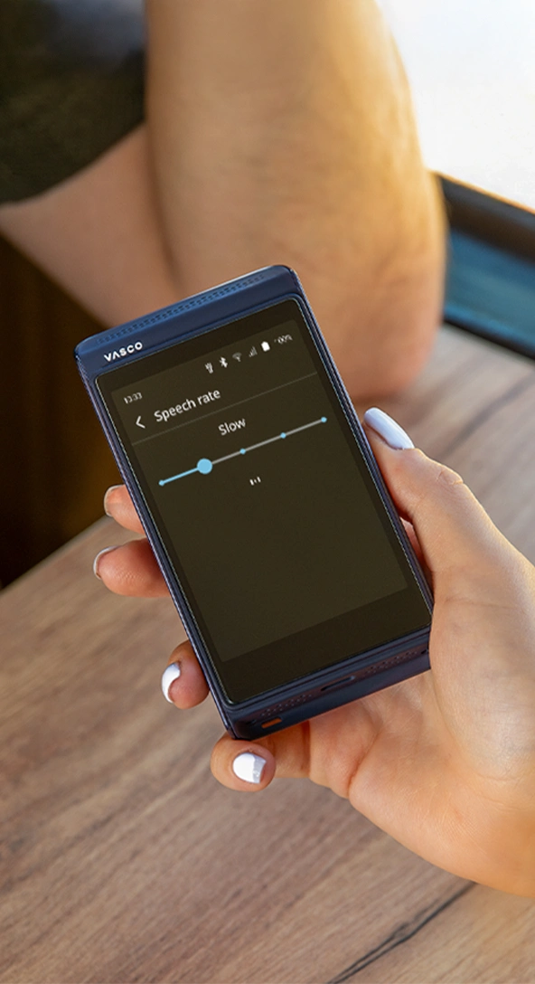 A hand holds a portable electronic device displaying a Preferred voices settings menu with options for user voice, conversation partner, and system voice. The person wears a yellow and white textured garment.