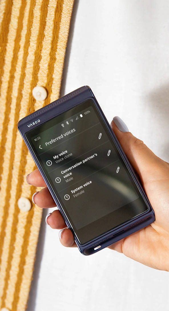 A person holds a VASCO electronic translator, displaying a screen with a Speech rate slider set to Slow. Another persons arm rests on a wooden table nearby.