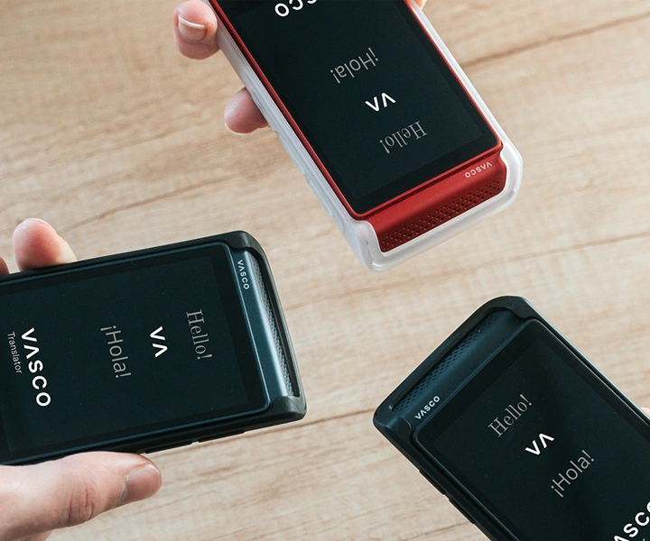 Three people holding Vasco Translator Q1 devices bring them together; the screens show the start screen with the Vasco logo and the words ‘Hello!’ and ‘¡Hola!’. The devices are presented in silicone cases in three colors: Solid Black, Dark Caramel, and Milky White.