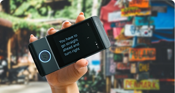 A close-up of a hand holding a black Vasco electronic translator horizontally. The screen is in full-screen mode, displaying the translated text in a large, clear blue font: 