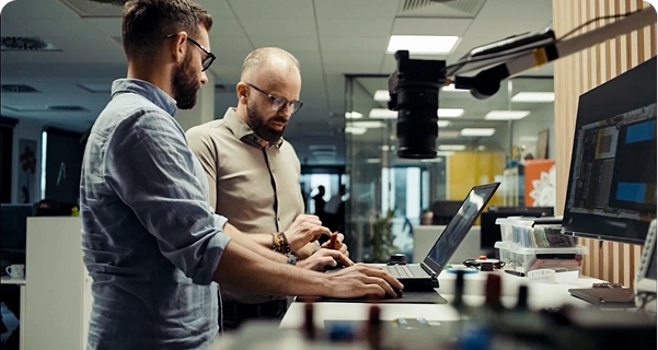 Two men with beards and glasses are working together at a standing desk in a modern office or laboratory. One man in a light blue shirt is using a laptop, while the other man in a tan shirt stands beside him, gesturing toward a small electronic component in his hand. The desk is equipped with a large monitor, various electronic tools, and a camera mounted on an adjustable arm. The background shows a bright, open-plan workspace with glass partitions. 