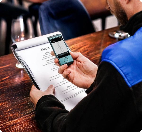 A close-up, high-angle shot of a person sitting at a wooden table in a restaurant, using a handheld electronic translator device to read a menu. The person holds a white paper menu in their left hand while holding a small, teal-colored Vasco translator over it with their right. The device’s screen shows a digital version of the menu being translated into Polish. The person is wearing a black jacket with a bright blue shoulder panel. The background is softly blurred, showing the warm interior of a dining establishment. 