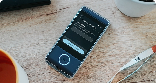 A high-angle, close-up shot of a dark blue Vasco electronic translator in a transparent protective case, resting on a light-colored wooden table. The device's screen is active, showing a 