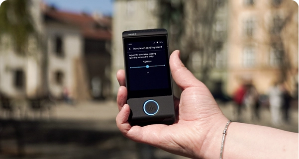 A close-up of a person's hand holding a black Vasco electronic translator against a blurred outdoor background of a city square with historic buildings. The device screen displays the 