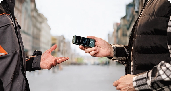 A person wearing a black quilted vest over a plaid shirt holds a teal Vasco electronic translator horizontally toward another person on a city street. The second person, wearing a grey and orange work jacket, holds their hand out as if mid-conversation. The device screen displays a translation interface with text. The background is a wide, blurred view of a European-style street with tall buildings under a bright, overcast sky. 