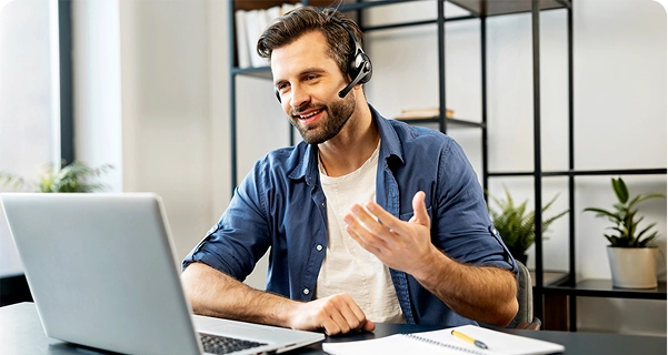 A friendly-looking man with a beard and dark hair sits at a desk, wearing a professional headset with a microphone. He is smiling and gesturing with one hand while looking at a laptop screen, appearing to be in the middle of a video call or providing technical support. He is dressed in a blue button-down shirt over a white t-shirt. On the desk in front of him are a laptop, a notebook, and a yellow pen. The background is a bright, modern office space with a minimalist black metal shelf and green potted plants. 