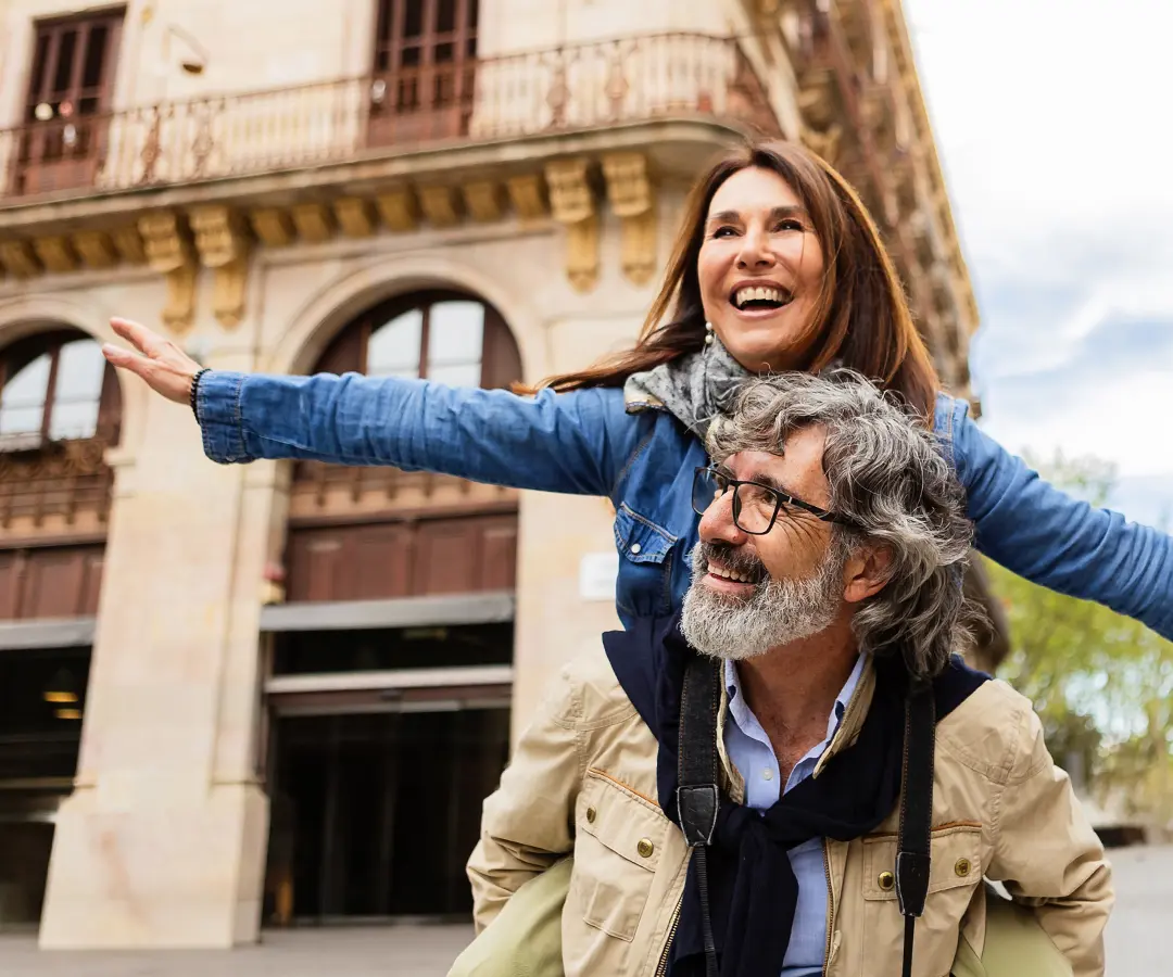 A joyful older man gives a woman a piggyback ride on a European city street, both smiling widely as she points with a raised arm toward the distance. He wears glasses, a tan jacket, and a camera, and she wears a denim jacket and scarf. 