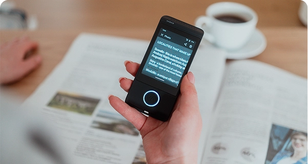A high-angle shot showing a person's hand holding a black Vasco electronic translator over an open magazine on a wooden table. The device's screen is in 
