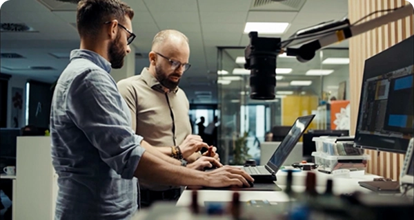 Two men with beards and glasses are working together at a standing desk in a modern office or laboratory. One man in a light blue shirt is using a laptop, while the other man in a tan shirt stands beside him, gesturing toward a small electronic component in his hand. The desk is equipped with a large monitor, various electronic tools, and a camera mounted on an adjustable arm. The background shows a bright, open-plan workspace with glass partitions.